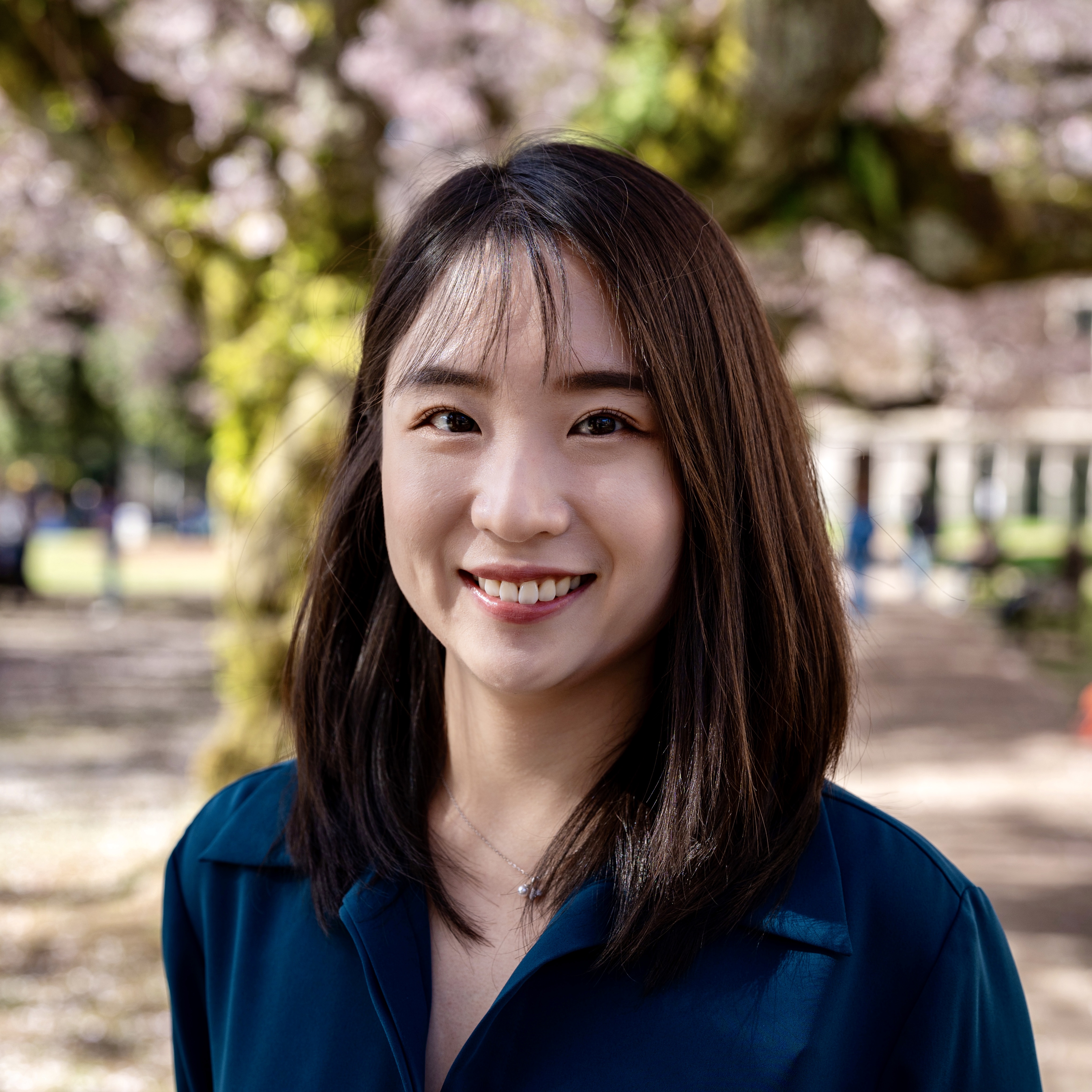 A picture of Chu, an asian woman with shoulder length brown hair. The background is cherry blossoms at the University of Washington.