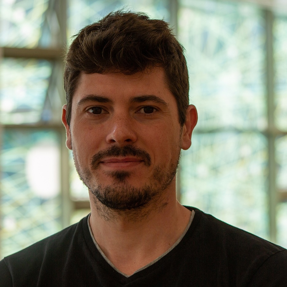 A headshot of Hugo Nicolau, a white man with brown short hair and beard, wearing a black shirt and stading in front of a glass window