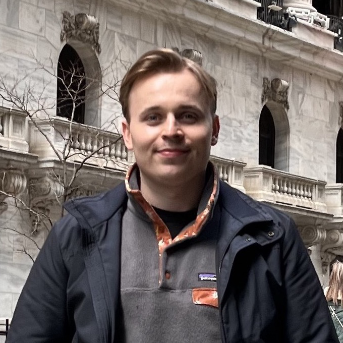 A picture of Humphrey, a white man with blonde hair, wearing a grey jumper, blue jacket t-shirt with grey Wall Street buildings as a background.