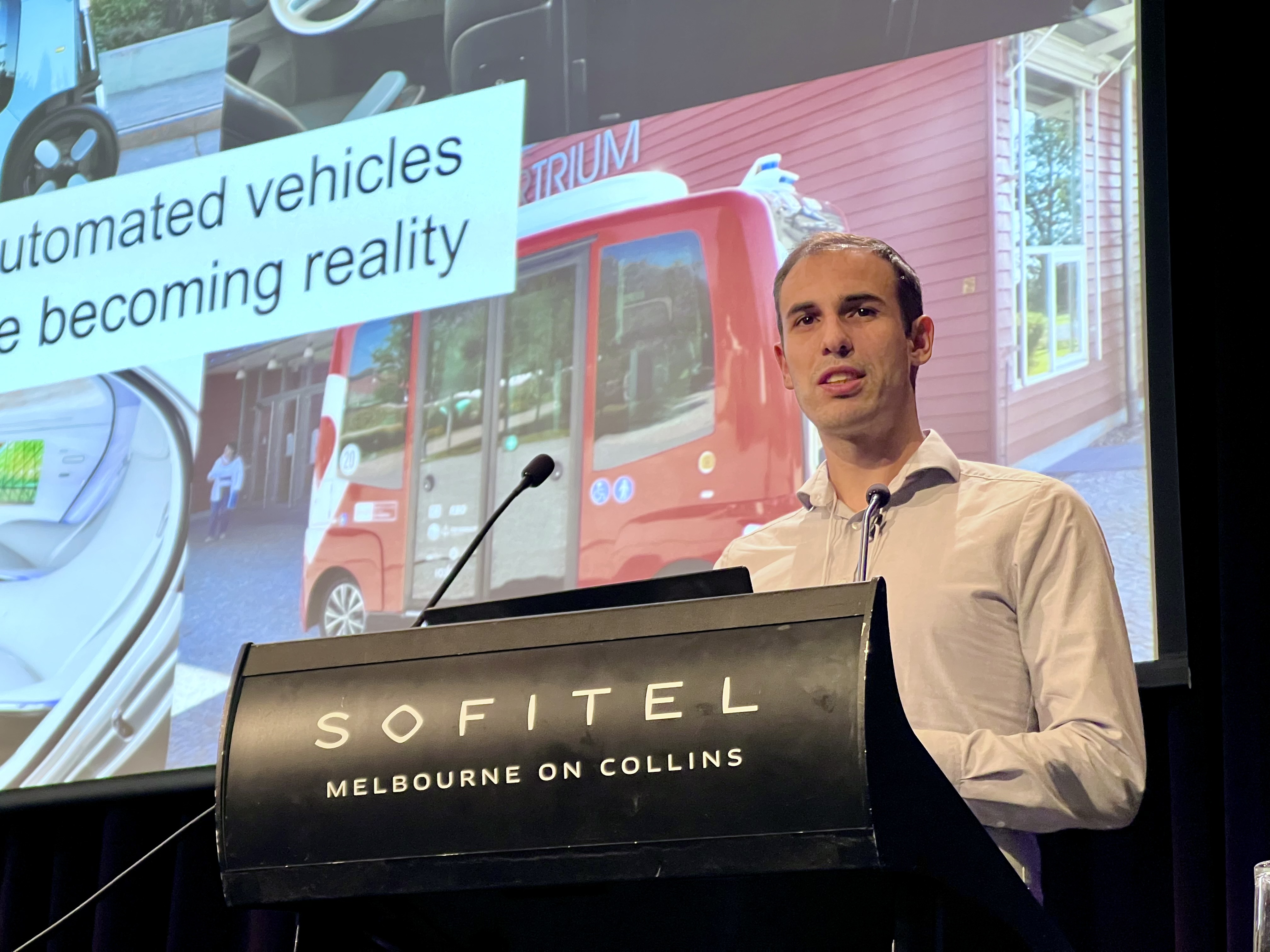 A portrait of Mark Colley. He is standing at a podium wearing a white shirt. He is giving a talk about automated vehicles, with slides about this topic presented in the background.