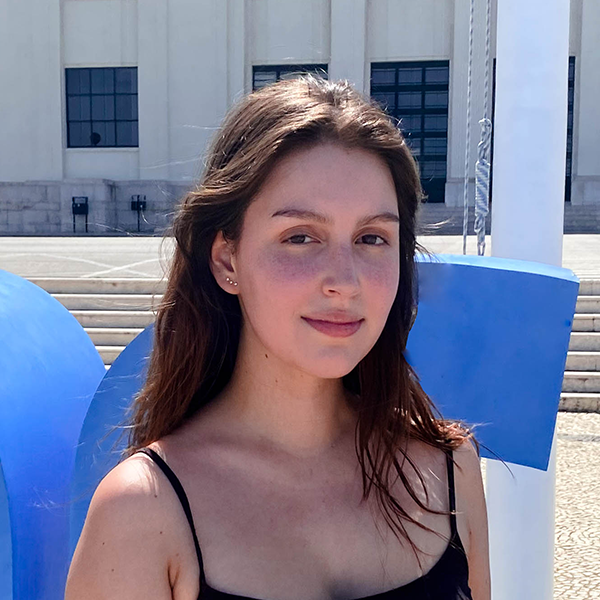 A headshot of Patricia Piedade, a white woman, she smiles softly, her cheeks are slightly red and freckled from the sun, her brown hair windswept, she wears a simple black summer top and stands in front of the engineering college building of the University of Lisbon.