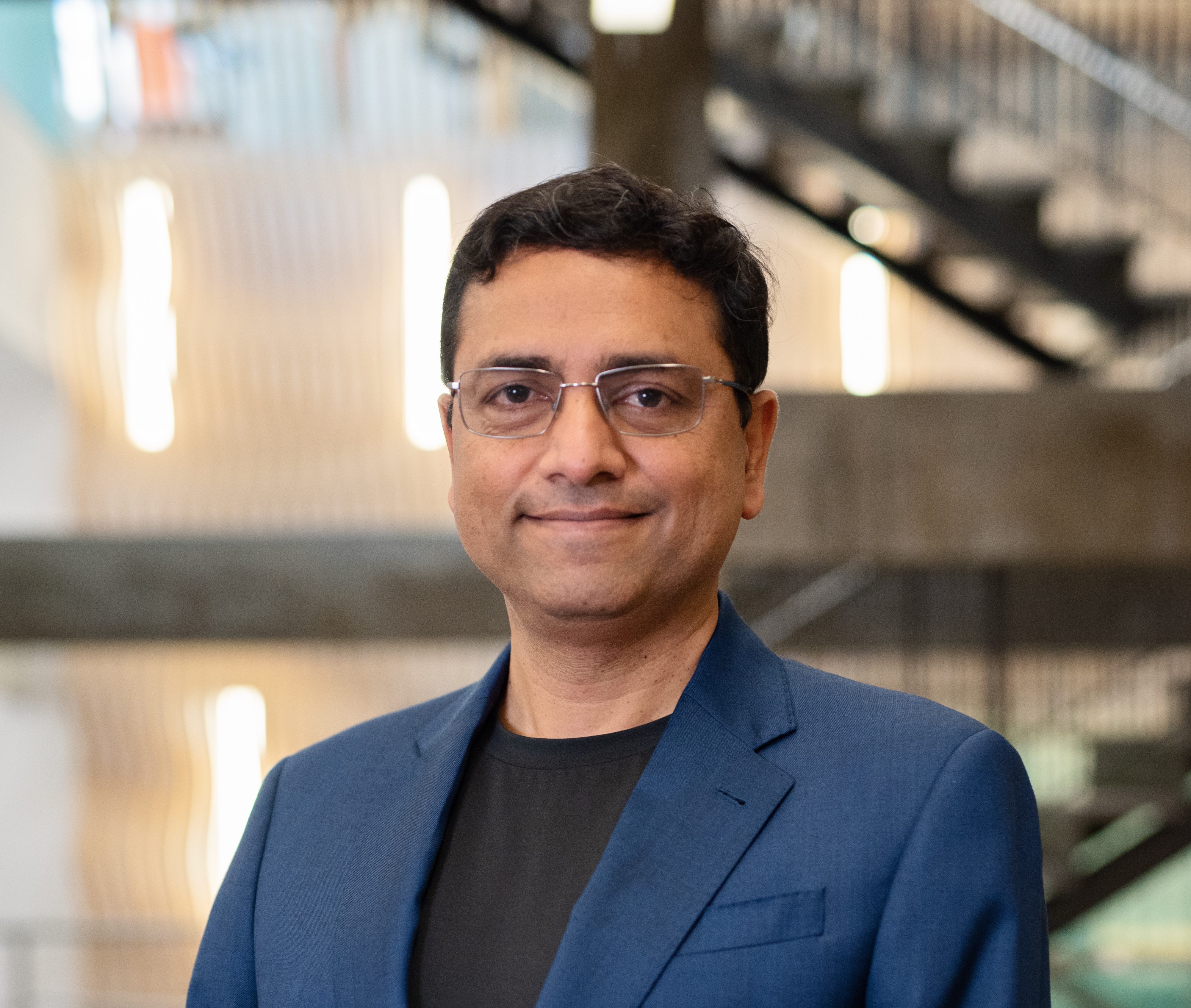 A picture of Raja, an Indian man with glasses, wearing a blue jacket over a black shirt, with a blurred background.